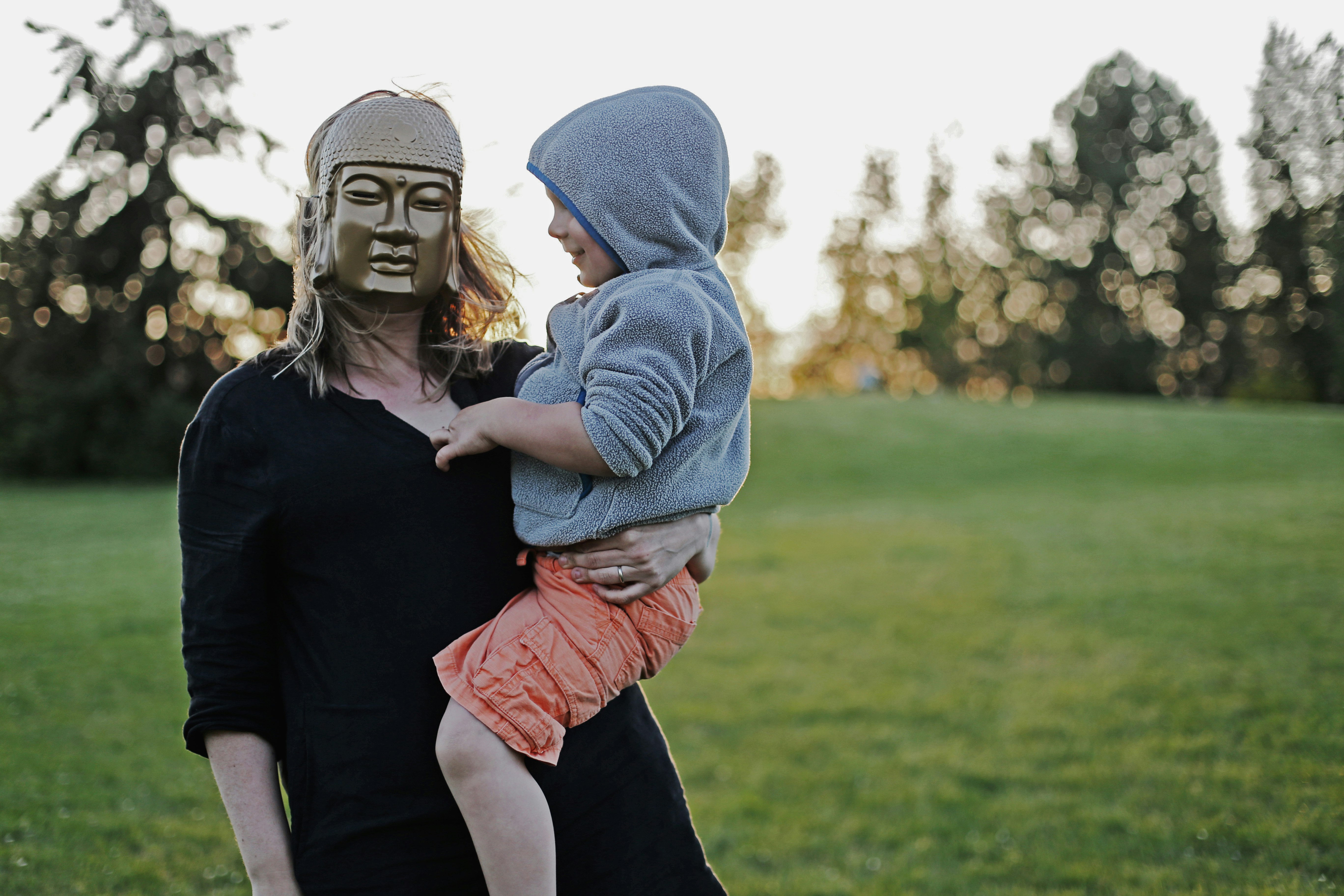 woman with mask carrying toddler with hoodie on grass field